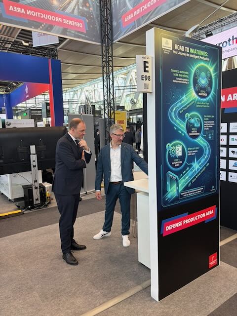 Zwei Gesprächspartner vor dem iniationWARE-Aufsteller „Road to iniationOS“ in der Messehalle der Hannover Messe 2026.