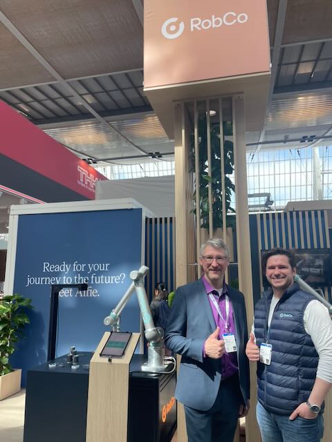 Zwei Personen mit Daumen hoch vor einem Cobot-Stand mit Roboterarm auf der Hannover Messe 2026.