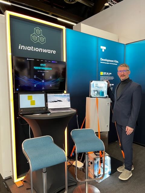 Mitarbeiter am iniationWARE-Stand der SPS Nürnberg 2025 vor Monitor mit OT/IT-Hybrid-Integrators und Demohardware.