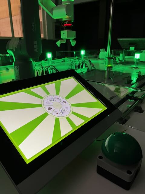 Tablet mit interaktiver Demo-Oberfläche und großer grüner Taste vor einem ABB-GoFa-Cobot am Messemodell 2023.