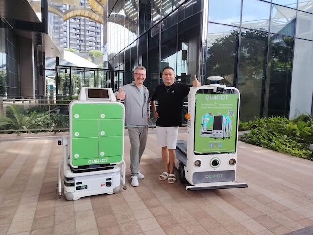 Zwei Personen zwischen zwei autonomen Lieferrobotern mit Fachfächern und Display im Außenbereich.